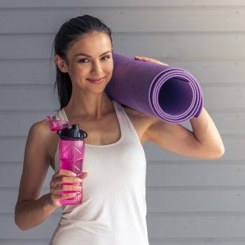 Beautiful young woman in sports wear is holding a yoga mat and a bottle of water, looking at camera and smiling, standing on a gray background